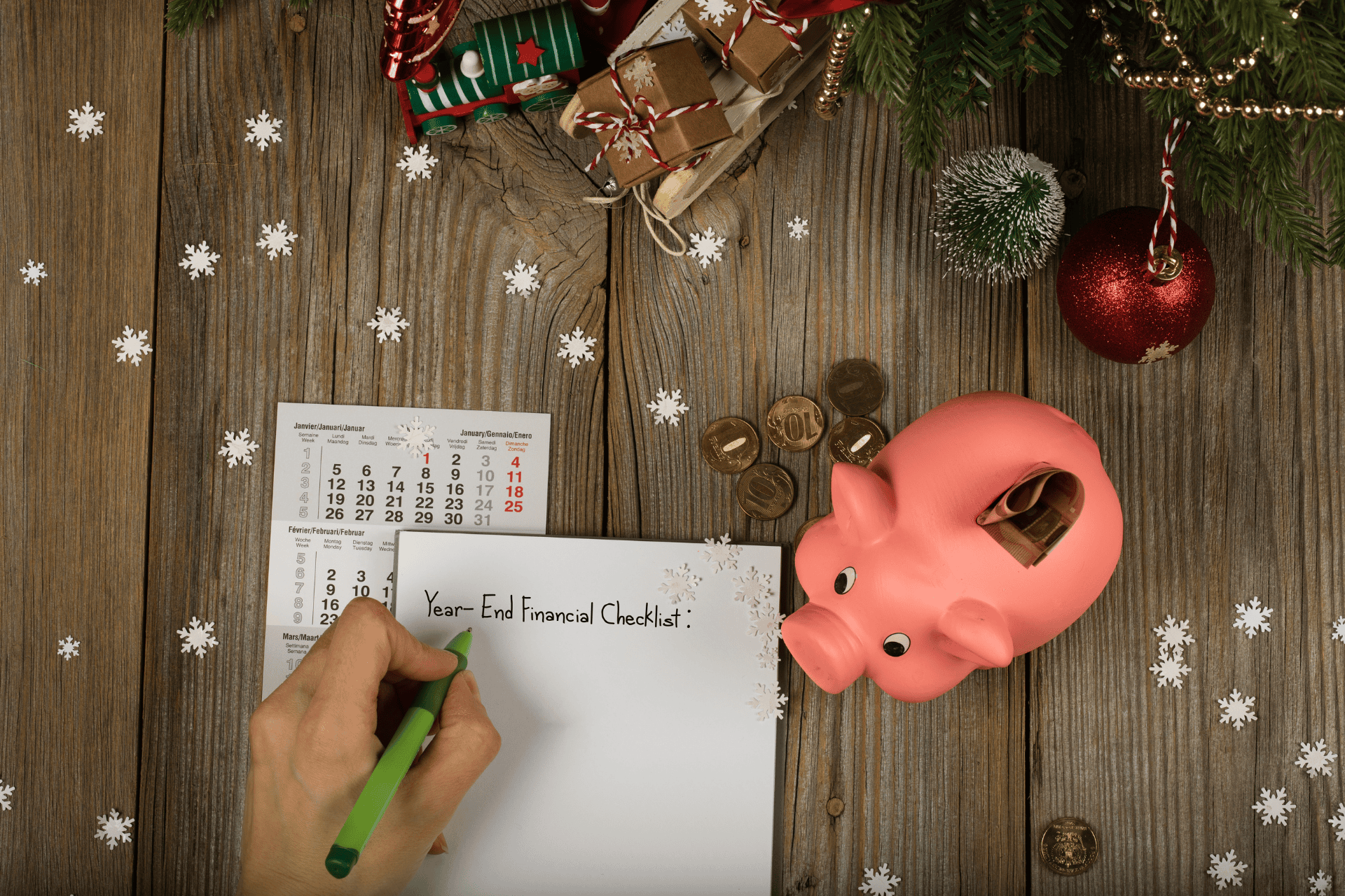 Hand writing "Year-End Financial Checklist" on a notepad surrounded by holiday decorations, a calendar, gold coins, and a pink piggy bank on a wooden table.